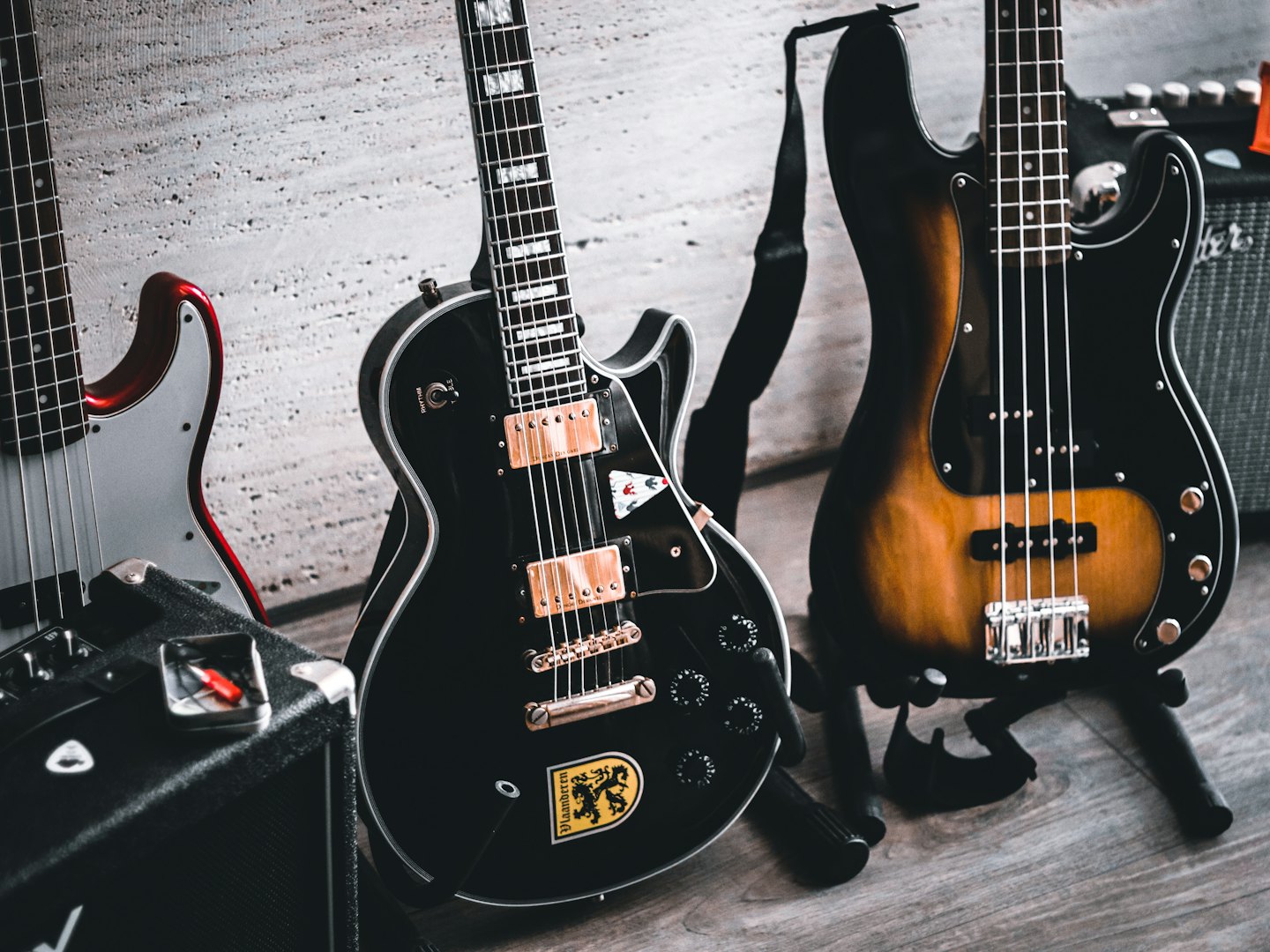 Three electric guitars and two amplifiers against a grey wall.